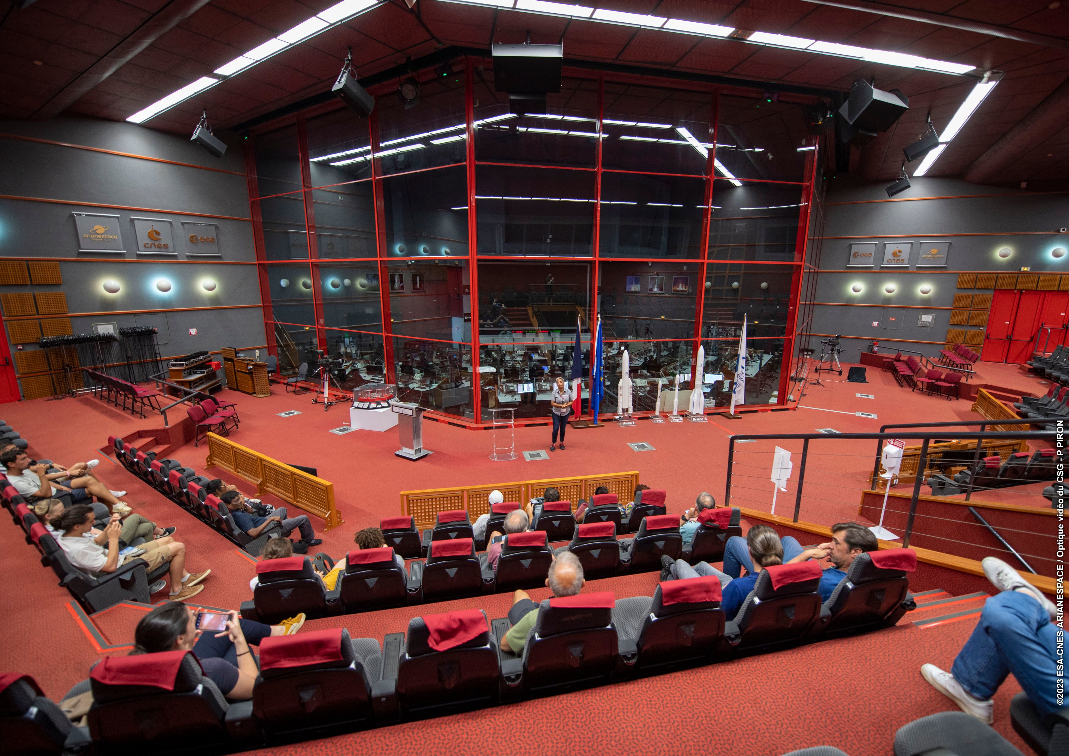 Une présentation se déroule actuellement dans la salle Jupiter, où des invités sont assis et écoutent un guide. En arrière-plan, on distingue la salle de contrôle.